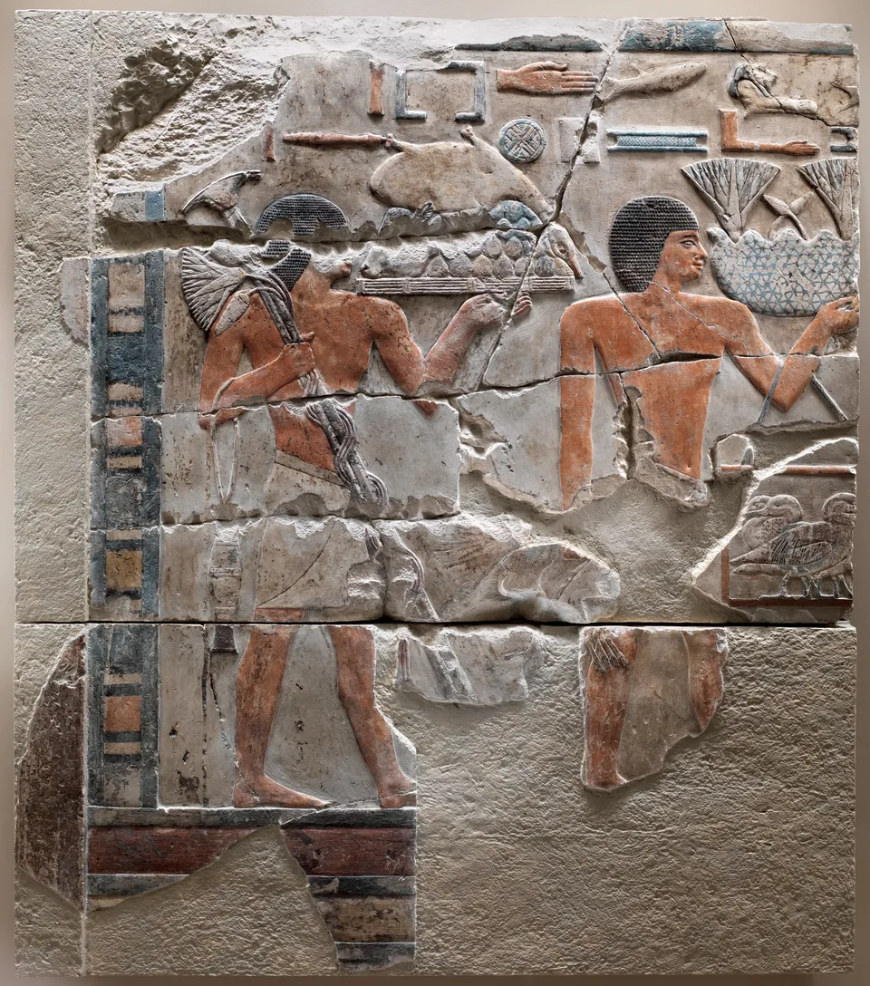 Two male offering bearers on joined limestone fragments, traces of red and blue paint, food tray at left, flowers and birds at right; inscriptions above.